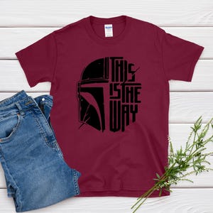 May include: Burgundy t-shirt with a black graphic of a helmet and the text "THIS IS THE WAY". The shirt is paired with blue jeans. The design is likely inspired by a popular science fiction franchise.