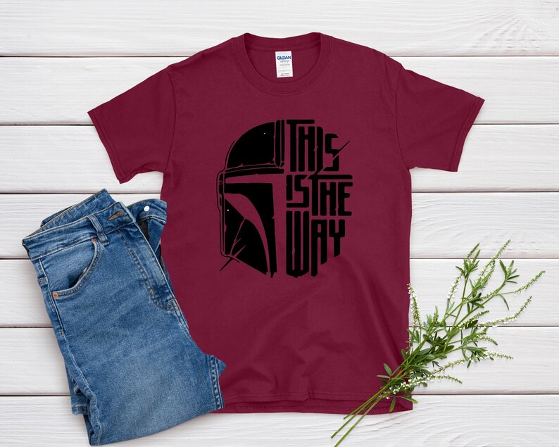 May include: Burgundy t-shirt with a black graphic of a helmet and the text "THIS IS THE WAY". The shirt is paired with blue jeans. The design is likely inspired by a popular science fiction franchise.