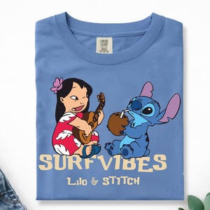 Puede incluir: Camiseta azul claro con Lilo y Stitch de Lilo & Stitch de Disney. Lilo toca el ukelele y Stitch bebe de un coco. Las palabras "SURF VIBES" y "Lilo & Stitch" est&aacute;n impresas en la camiseta.