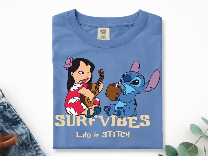 Puede incluir: Camiseta azul claro con Lilo y Stitch de Lilo & Stitch de Disney. Lilo toca el ukelele y Stitch bebe de un coco. Las palabras "SURF VIBES" y "Lilo & Stitch" est&aacute;n impresas en la camiseta.