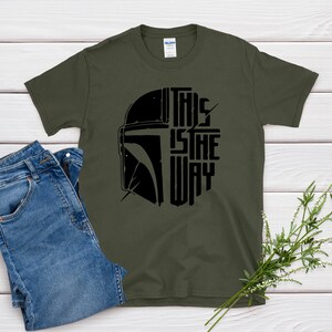 May include: Olive green t-shirt with a black graphic of a helmet and the text "THIS IS THE WAY." The shirt is paired with blue jeans. A sprig of greenery is placed on the right side of the image.