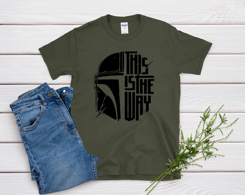 May include: Olive green t-shirt with a black graphic of a helmet and the text "THIS IS THE WAY." The shirt is paired with blue jeans. A sprig of greenery is placed on the right side of the image.