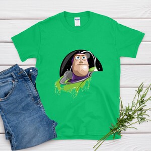May include: A bright green t-shirt featuring a cartoon graphic of Buzz Lightyear from Toy Story. The graphic shows Buzz in his space suit against a starry background. Blue denim jeans are next to the shirt on a white wooden surface.