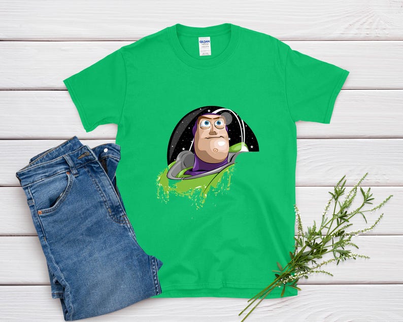 May include: A bright green t-shirt featuring a cartoon graphic of Buzz Lightyear from Toy Story. The graphic shows Buzz in his space suit against a starry background. Blue denim jeans are next to the shirt on a white wooden surface.