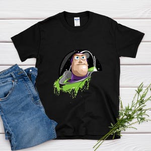 May include: Black t-shirt featuring a graphic of Buzz Lightyear from Toy Story. The design shows Buzz in his space suit, with a green and purple color scheme. The shirt is paired with blue jeans on a white wooden surface.