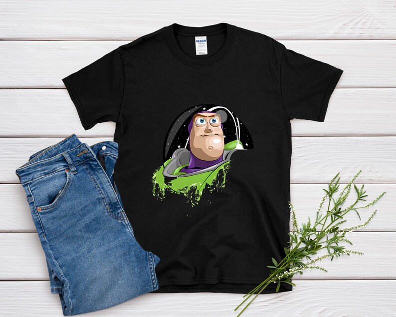May include: Black t-shirt featuring a graphic of Buzz Lightyear from Toy Story. The design shows Buzz in his space suit, with a green and purple color scheme. The shirt is paired with blue jeans on a white wooden surface.