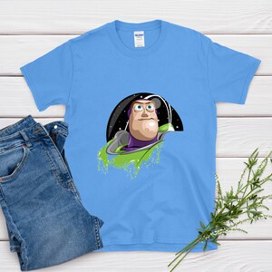 May include: A light blue t-shirt featuring a graphic of Buzz Lightyear from Toy Story. The design shows Buzz in his space suit against a black and starry background. A pair of blue jeans is next to the shirt.