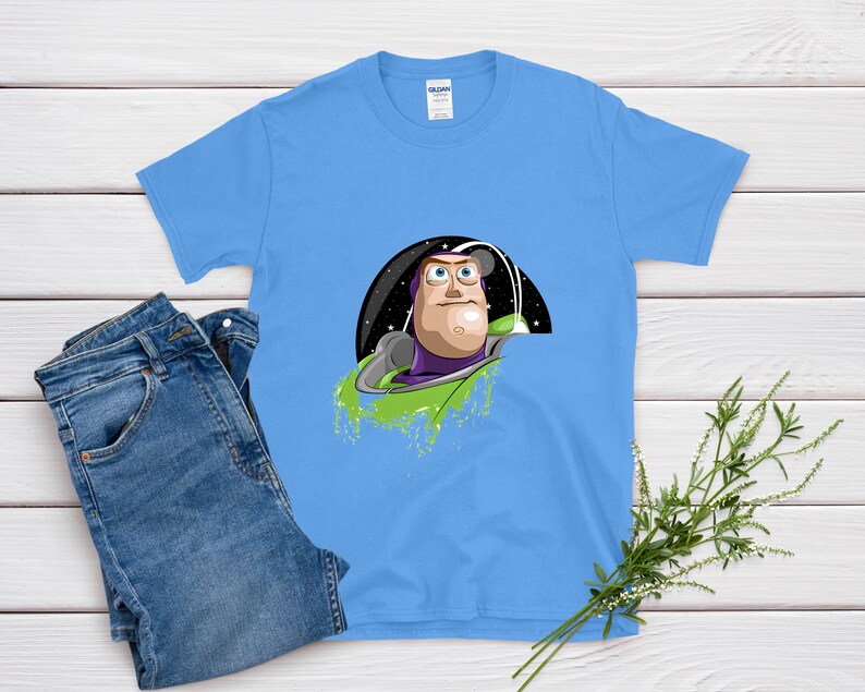 May include: A light blue t-shirt featuring a graphic of Buzz Lightyear from Toy Story. The design shows Buzz in his space suit against a black and starry background. A pair of blue jeans is next to the shirt.