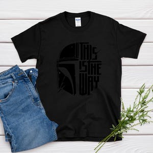 May include: Black t-shirt with a graphic of a helmet and the text "THIS IS THE WAY" in a stylized font. The shirt is paired with blue denim jeans. The design is centered on the shirt.