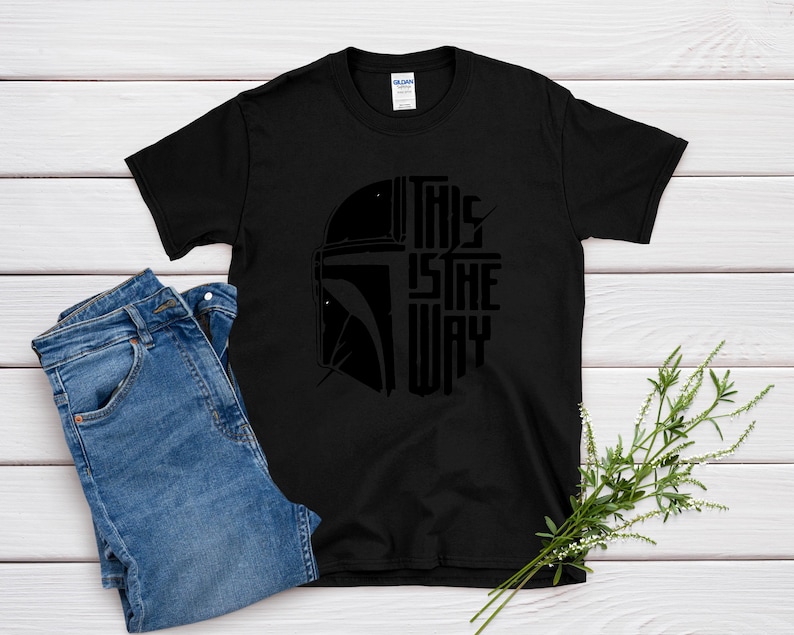 May include: Black t-shirt with a graphic of a helmet and the text "THIS IS THE WAY" in a stylized font. The shirt is paired with blue denim jeans. The design is centered on the shirt.