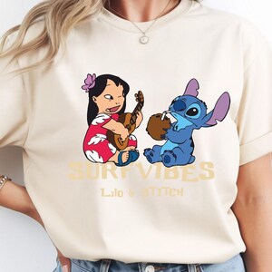 Puede incluir: Camiseta beige con un dise&ntilde;o de Lilo tocando el ukelele y Stitch bebiendo de un coco. Las palabras "SURF VIBES" y "Lilo & Stitch" est&aacute;n impresas en dorado.