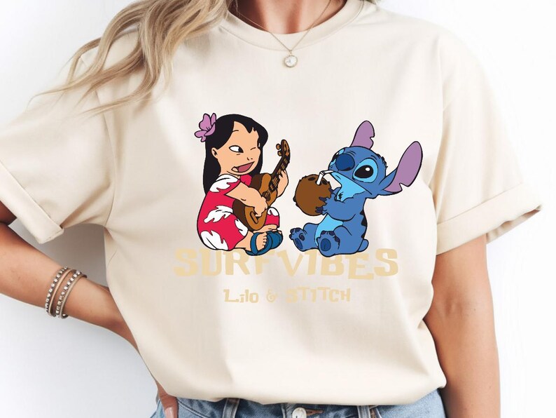 Puede incluir: Camiseta beige con un dise&ntilde;o de Lilo tocando el ukelele y Stitch bebiendo de un coco. Las palabras "SURF VIBES" y "Lilo & Stitch" est&aacute;n impresas en dorado.