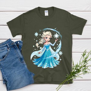 May include: An olive green t-shirt featuring a cartoon illustration of a princess in a blue dress with snowflakes. The princess has blonde hair and is surrounded by sparkling stars and a blue arc. A pair of blue jeans is visible on the left.