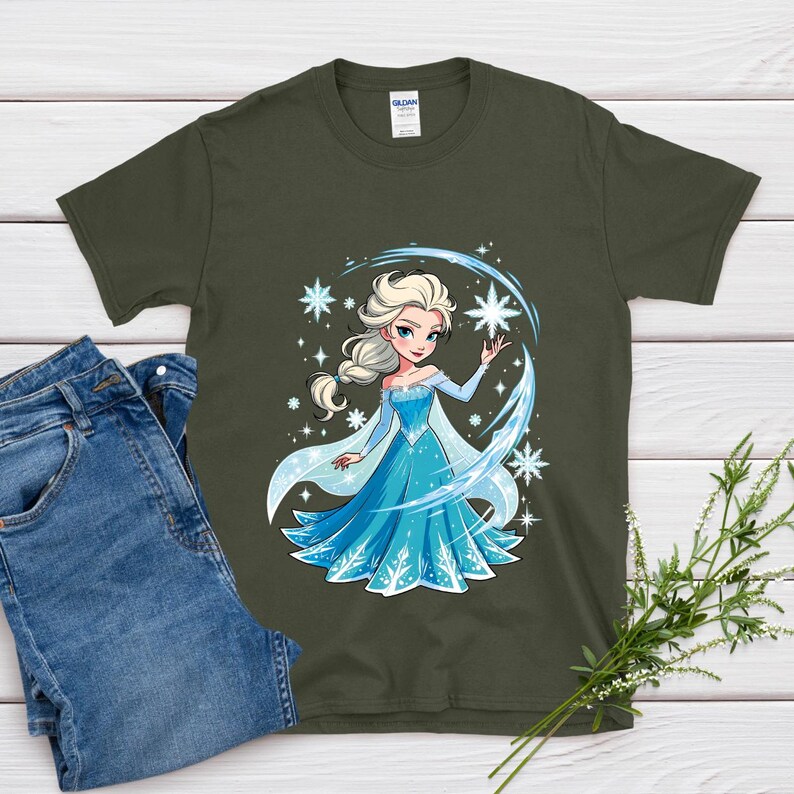 May include: An olive green t-shirt featuring a cartoon illustration of a princess in a blue dress with snowflakes. The princess has blonde hair and is surrounded by sparkling stars and a blue arc. A pair of blue jeans is visible on the left.