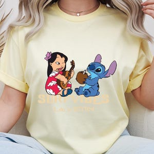 Puede incluir: Una camiseta amarilla clara con un gr&aacute;fico de Lilo tocando el ukelele y Stitch bebiendo de un coco. El texto "SURF VIBES" y "Life is better" est&aacute; impreso debajo. La camiseta tiene mangas enrolladas.