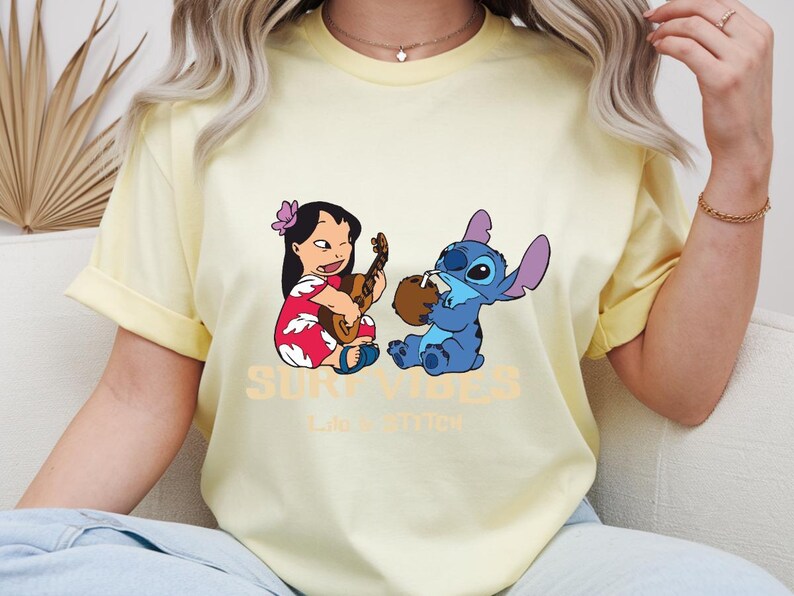 Puede incluir: Una camiseta amarilla clara con un gr&aacute;fico de Lilo tocando el ukelele y Stitch bebiendo de un coco. El texto "SURF VIBES" y "Life is better" est&aacute; impreso debajo. La camiseta tiene mangas enrolladas.