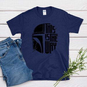May include: Navy blue t-shirt featuring a black graphic of a helmet and the text "THIS IS THE WAY". The shirt is paired with blue denim jeans. A sprig of greenery is placed on the side.