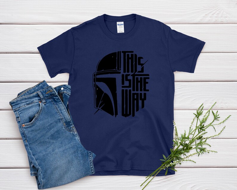 May include: Navy blue t-shirt featuring a black graphic of a helmet and the text "THIS IS THE WAY". The shirt is paired with blue denim jeans. A sprig of greenery is placed on the side.