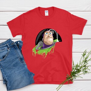 May include: A red t-shirt featuring a graphic of Buzz Lightyear from Toy Story. The design shows Buzz in his space suit, set against a black and starry background with green accents. Blue jeans are next to the shirt.