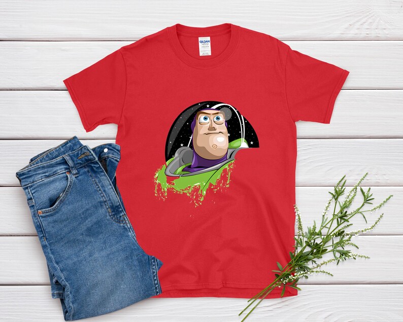 May include: A red t-shirt featuring a graphic of Buzz Lightyear from Toy Story. The design shows Buzz in his space suit, set against a black and starry background with green accents. Blue jeans are next to the shirt.