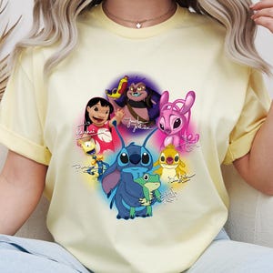 Op de afbeelding: Een lichtgeel T-shirt met een kleurrijke afbeelding van personages uit de animatiefilm Lilo & Stitch. Het ontwerp bevat Stitch, Lilo en andere personages met hun namen eronder geschreven. Het shirt heeft korte mouwen.
