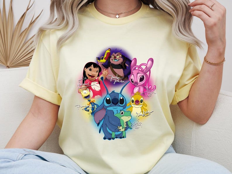 Op de afbeelding: Een lichtgeel T-shirt met een kleurrijke afbeelding van personages uit de animatiefilm Lilo & Stitch. Het ontwerp bevat Stitch, Lilo en andere personages met hun namen eronder geschreven. Het shirt heeft korte mouwen.