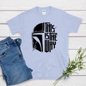 May include: Light blue t-shirt with a black graphic of a helmet and the text "THIS IS THE WAY". The shirt is paired with blue jeans. A sprig of greenery is placed on a white wooden surface.