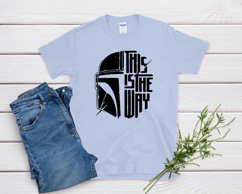 May include: Light blue t-shirt with a black graphic of a helmet and the text "THIS IS THE WAY". The shirt is paired with blue jeans. A sprig of greenery is placed on a white wooden surface.