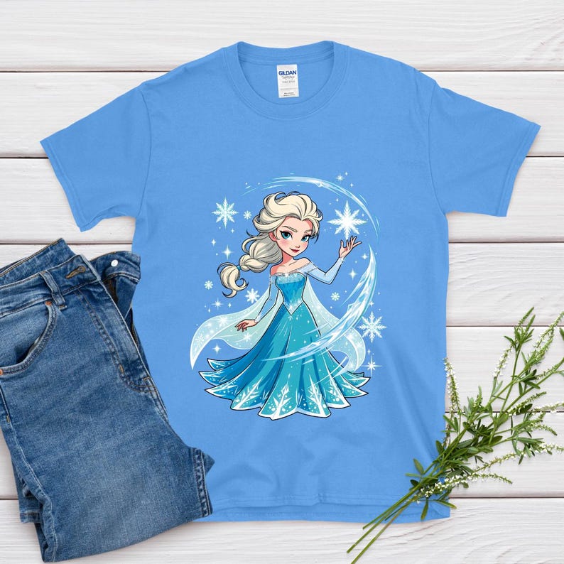 May include: A light blue t-shirt featuring a cartoon illustration of a princess in a blue dress with snowflakes. The princess has blonde hair and is surrounded by a crescent of snowflakes. A pair of blue jeans is next to the shirt.