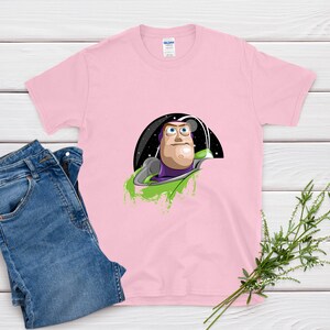 May include: A light pink t-shirt featuring a graphic of Buzz Lightyear from Toy Story. The design shows Buzz in his space suit, set against a black and starry background with green accents. Blue jeans are folded next to the shirt.