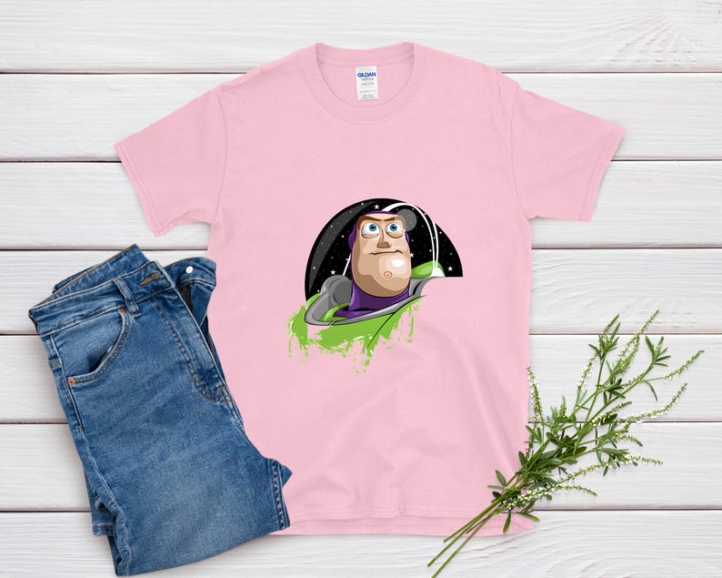 May include: A light pink t-shirt featuring a graphic of Buzz Lightyear from Toy Story. The design shows Buzz in his space suit, set against a black and starry background with green accents. Blue jeans are folded next to the shirt.