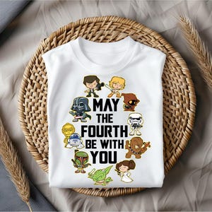 May include: White t-shirt featuring cartoon characters from Star Wars. The shirt has the text "May the Fourth Be With You" in black. The characters are arranged in a circle around the text.