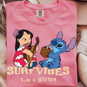 Puede incluir: Camiseta rosa Comfort Colors con un gr&aacute;fico de Lilo tocando el ukelele y Stitch bebiendo de un coco. Las palabras "SURF VIBES" y "Lilo & Stitch" est&aacute;n impresas en amarillo claro.
