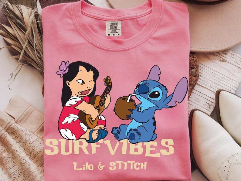 Puede incluir: Camiseta rosa Comfort Colors con un gr&aacute;fico de Lilo tocando el ukelele y Stitch bebiendo de un coco. Las palabras "SURF VIBES" y "Lilo & Stitch" est&aacute;n impresas en amarillo claro.