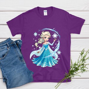 May include: A purple t-shirt featuring a cartoon illustration of a princess in a blue dress with a white cape, surrounded by snowflakes and a crescent moon design. The shirt is paired with blue jeans.