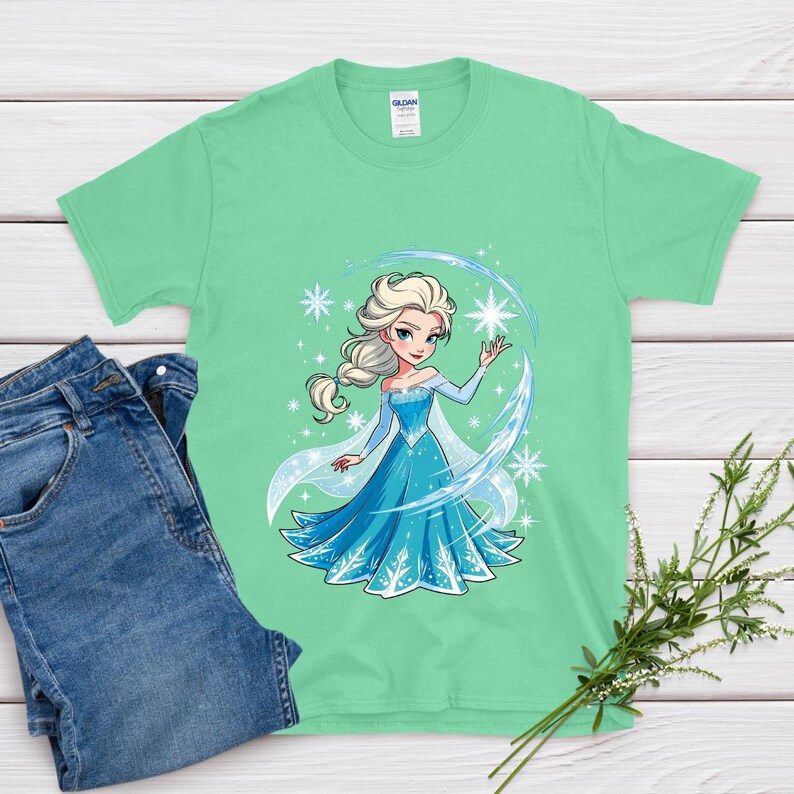 May include: A mint green t-shirt featuring a cartoon illustration of a princess in a blue dress with snowflakes. The princess has blonde hair and is surrounded by a blue arc and stars. A pair of blue jeans is next to the shirt.