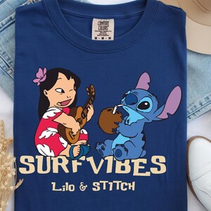 Puede incluir: Camiseta azul marino con una ilustraci&oacute;n de dibujos animados de Lilo tocando el ukelele y Stitch bebiendo de un coco. Las palabras "SURF VIBES" y "Lilo & Stitch" est&aacute;n impresas en amarillo.