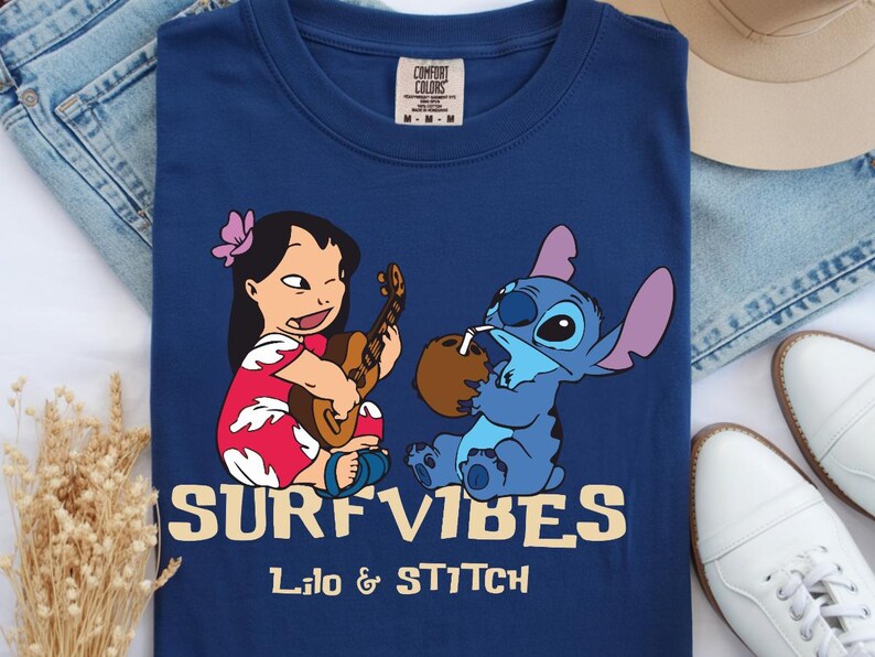 Puede incluir: Camiseta azul marino con una ilustraci&oacute;n de dibujos animados de Lilo tocando el ukelele y Stitch bebiendo de un coco. Las palabras "SURF VIBES" y "Lilo & Stitch" est&aacute;n impresas en amarillo.