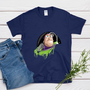 May include: Navy blue t-shirt featuring a cartoon graphic of Buzz Lightyear from Toy Story. The graphic shows Buzz in his space suit, with a green splash effect. A pair of blue jeans is next to the shirt.