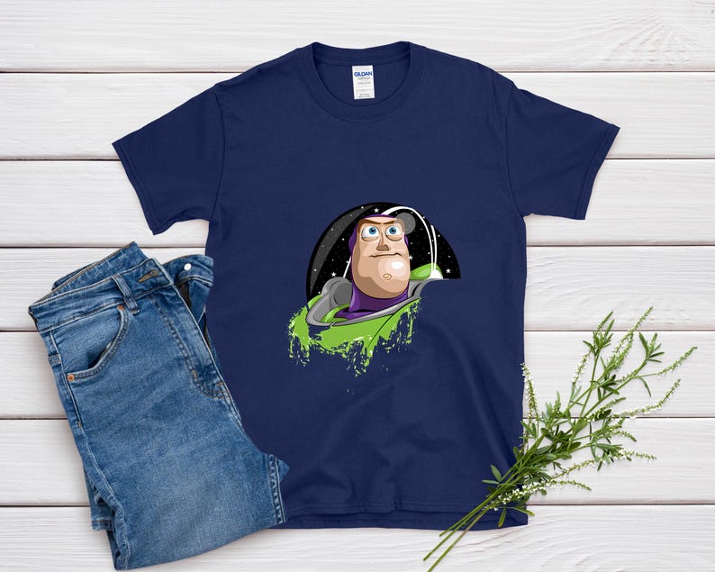 May include: Navy blue t-shirt featuring a cartoon graphic of Buzz Lightyear from Toy Story. The graphic shows Buzz in his space suit, with a green splash effect. A pair of blue jeans is next to the shirt.