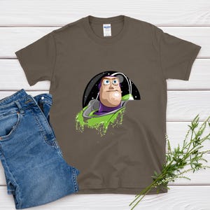 May include: A brown t-shirt featuring a cartoon image of Buzz Lightyear in his spaceship. The graphic includes a black space background and green accents. Blue jeans are folded next to the shirt on a white wooden surface.