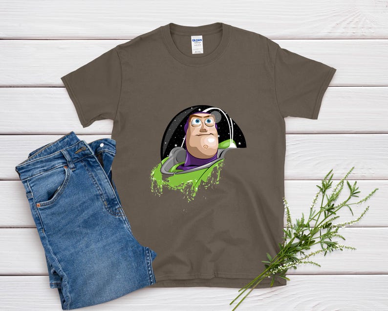 May include: A brown t-shirt featuring a cartoon image of Buzz Lightyear in his spaceship. The graphic includes a black space background and green accents. Blue jeans are folded next to the shirt on a white wooden surface.