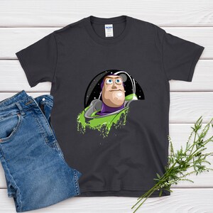 May include: Dark gray t-shirt featuring a graphic of Buzz Lightyear from Toy Story. The design shows Buzz in his space suit against a starry background with green accents. A pair of blue jeans is next to the shirt.