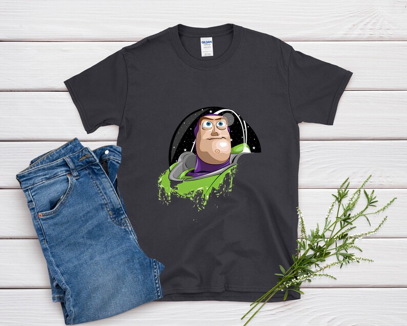 May include: Dark gray t-shirt featuring a graphic of Buzz Lightyear from Toy Story. The design shows Buzz in his space suit against a starry background with green accents. A pair of blue jeans is next to the shirt.
