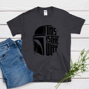 May include: Dark gray t-shirt with a black graphic of a helmet and the text "THIS IS THE WAY." The shirt is paired with blue jeans. A sprig of greenery is placed on the side.