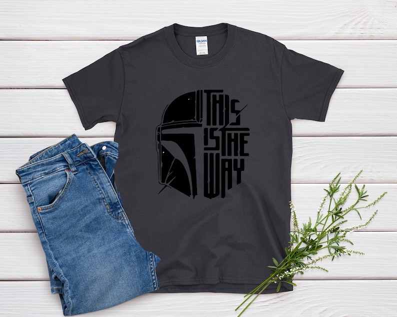 May include: Dark gray t-shirt with a black graphic of a helmet and the text "THIS IS THE WAY." The shirt is paired with blue jeans. A sprig of greenery is placed on the side.
