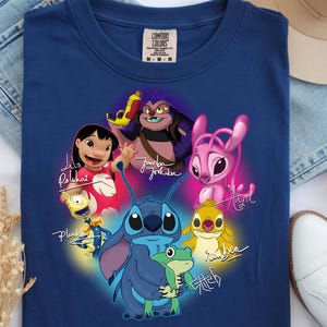 Op de afbeelding: Een marineblauw t-shirt met een kleurrijke afbeelding van personages uit de animatiefilm "Lilo & Stitch". Het ontwerp bevat Stitch, Lilo en andere personages met hun namen in een gestileerd lettertype. Het shirt heeft een ronde hals en korte mouwen.