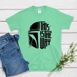 May include: A light green t-shirt with a black graphic of a helmet and the text "THIS IS THE WAY". The shirt is paired with blue jeans. A sprig of greenery is in the bottom right corner.