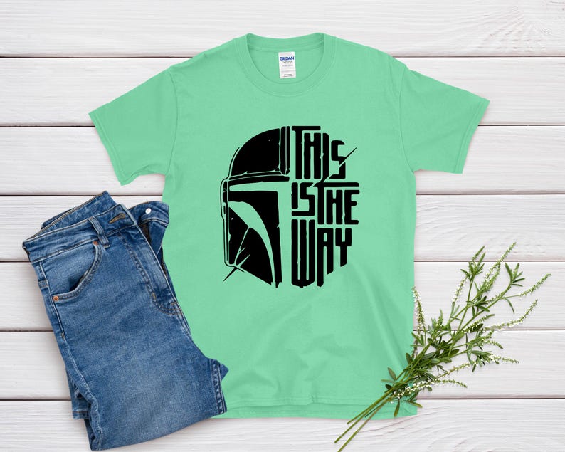 May include: A light green t-shirt with a black graphic of a helmet and the text "THIS IS THE WAY". The shirt is paired with blue jeans. A sprig of greenery is in the bottom right corner.