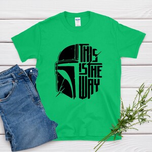 May include: A green t-shirt with a black graphic of a helmet and the text "THIS IS THE WAY." The shirt is paired with blue jeans. A sprig of greenery rests on the wooden surface.